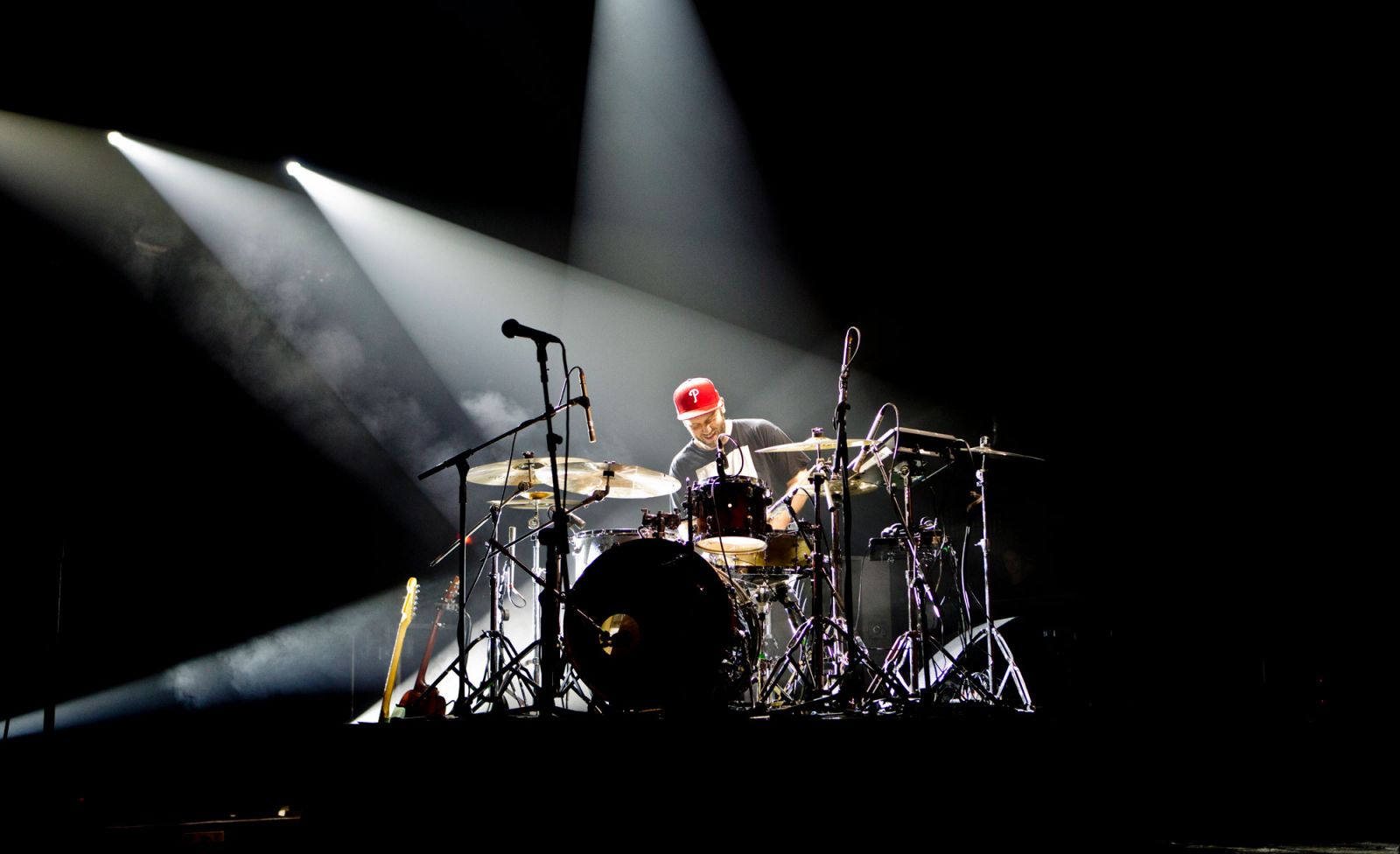 Drummer in a red cap playing a kit on a darkened stage, lit by two crossing white spotlights through haze